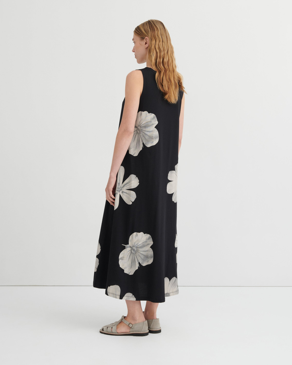 A woman stands with her back to the camera wearing a sleeveless black midi dress with a large white floral print, paired with gray strappy flat sandals, against a plain white studio background.