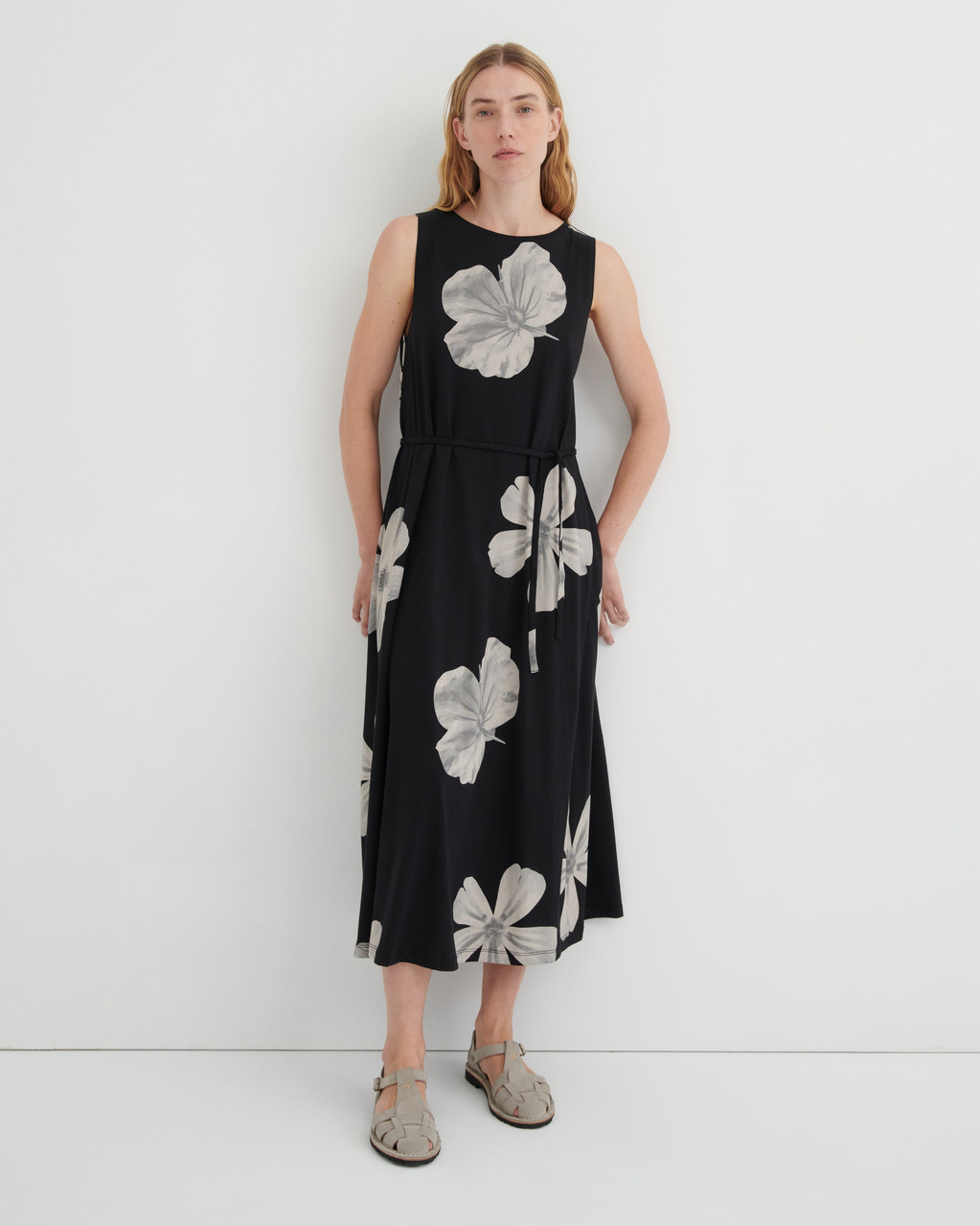 A woman modeling a sleeveless black midi dress with a large gray floral print, standing against a plain white studio background.