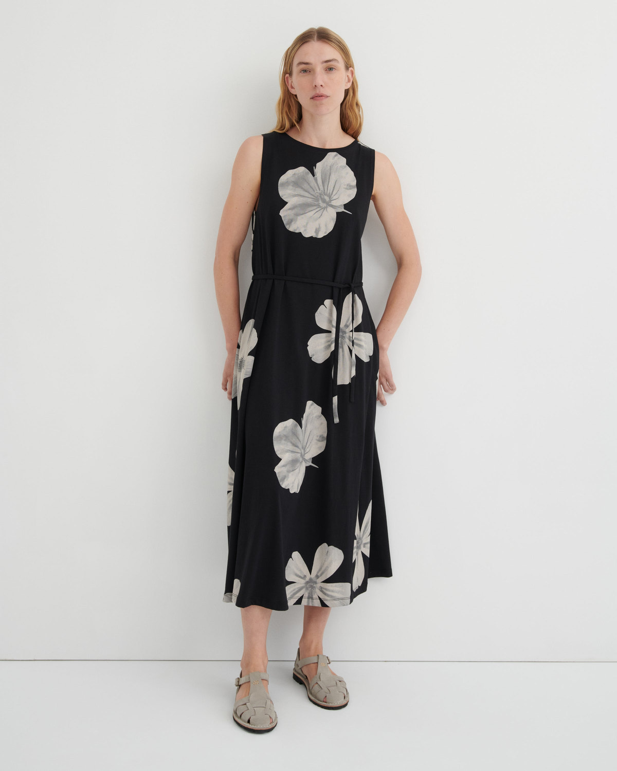 A woman modeling a sleeveless black midi dress with a large gray floral print, standing against a plain white studio background.
