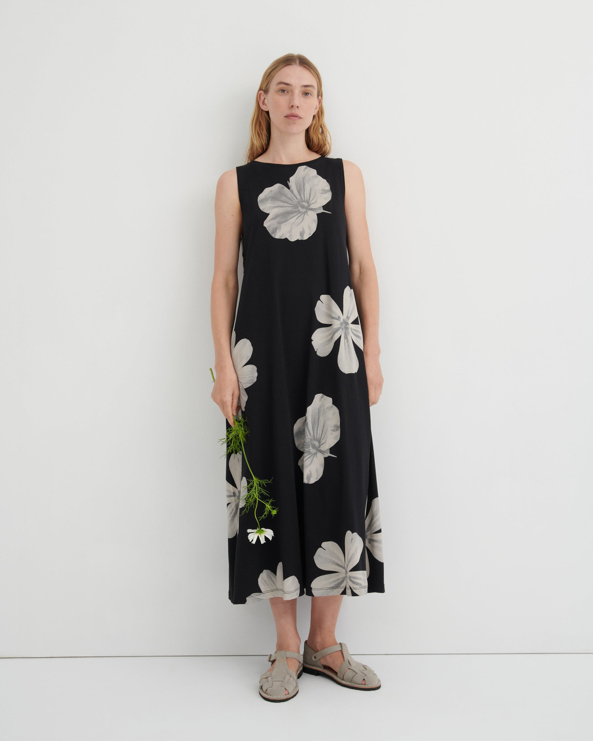 A woman stands against a plain white studio wall wearing a sleeveless black midi dress with a large white floral print. She is holding a small green sprig and wearing flat tan sandals.