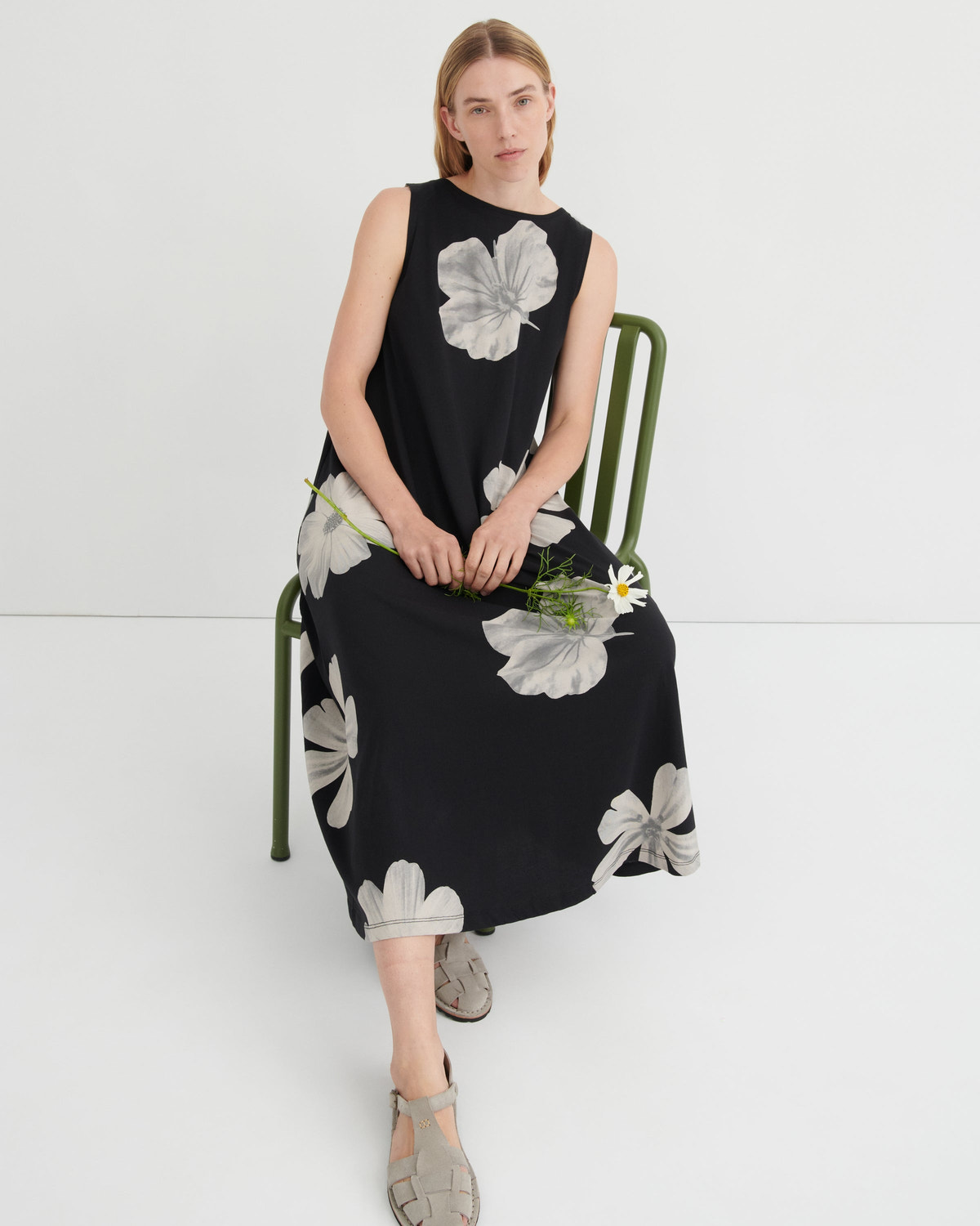 A woman sits on a green metal chair wearing a sleeveless black midi dress with a large white floral print. She is holding a small white flower and wearing gray strappy sandals against a clean white studio background.