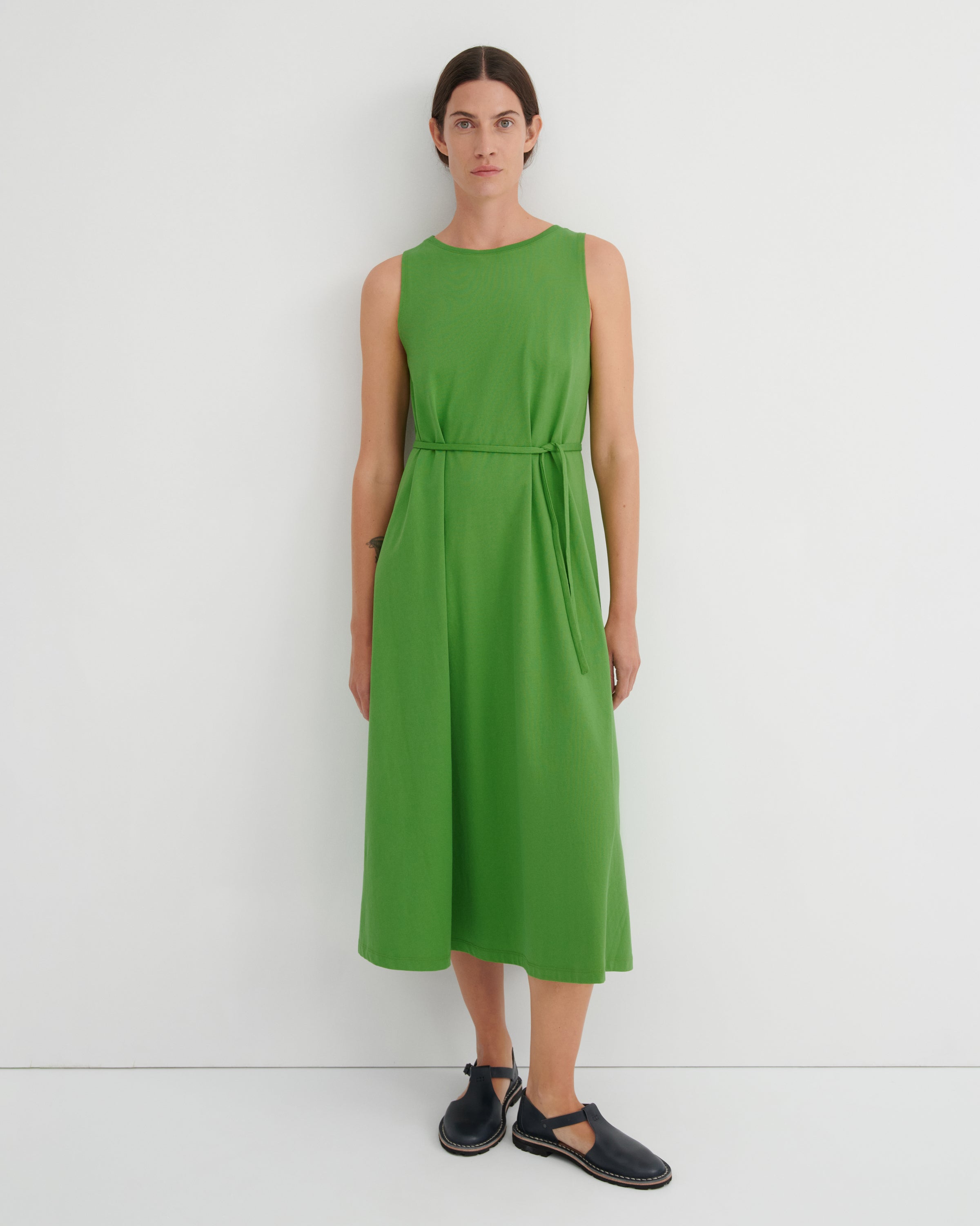 A woman stands against a plain white wall wearing a sleeveless bright green midi dress with a tie waist and dark navy flat shoes.