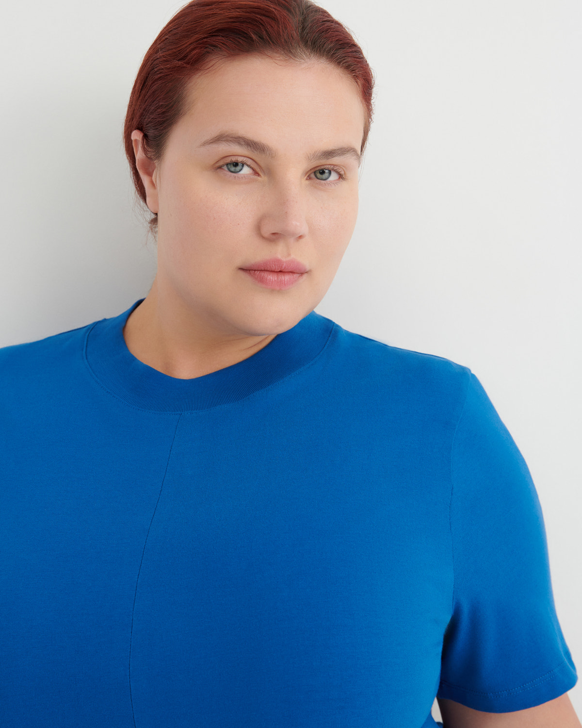 A close-up shot of a woman with red hair wearing a bright blue short-sleeve crewneck top against a light gray background.