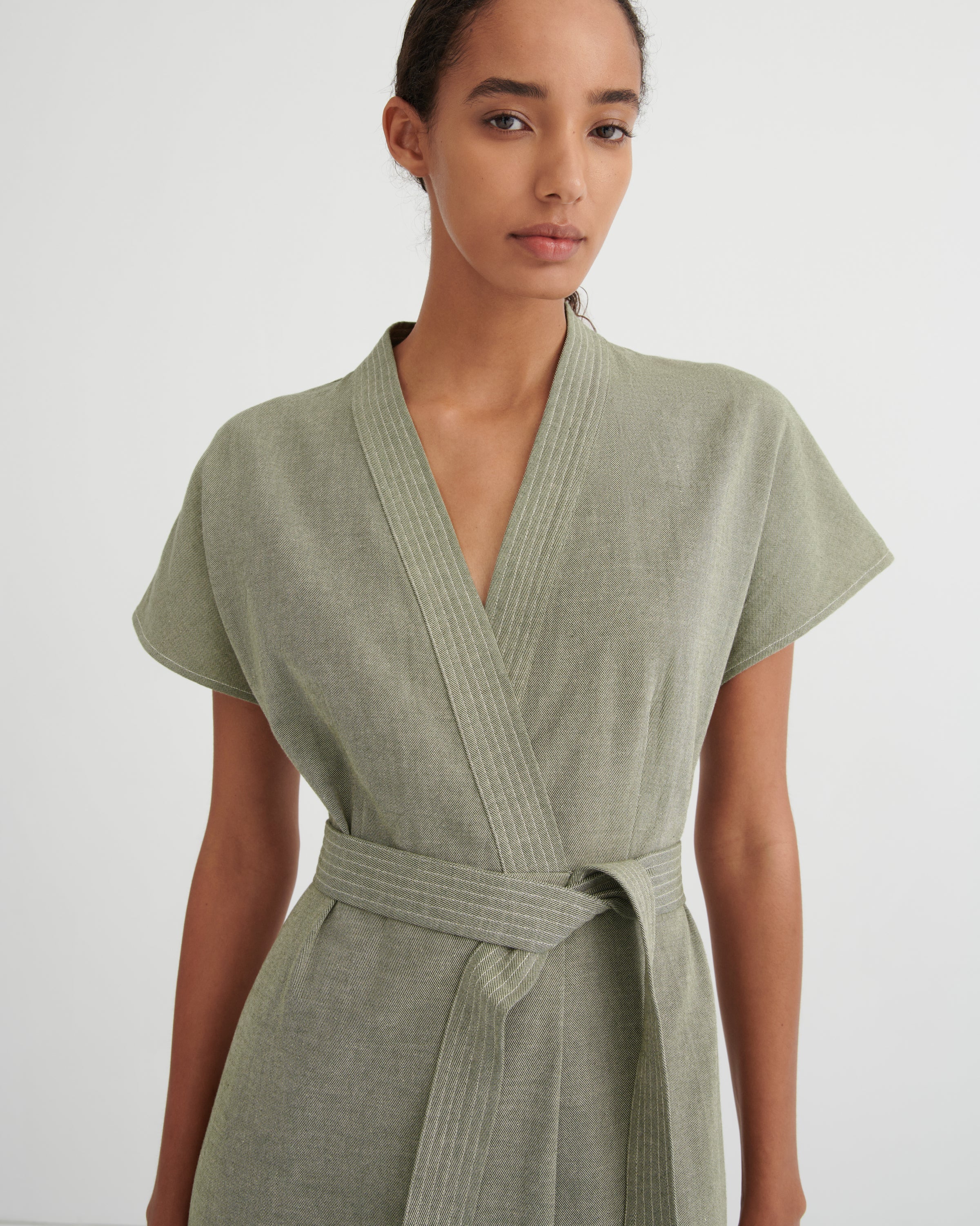 A woman wearing a sage green short-sleeve wrap dress with a matching tie belt, photographed against a plain light gray studio background.