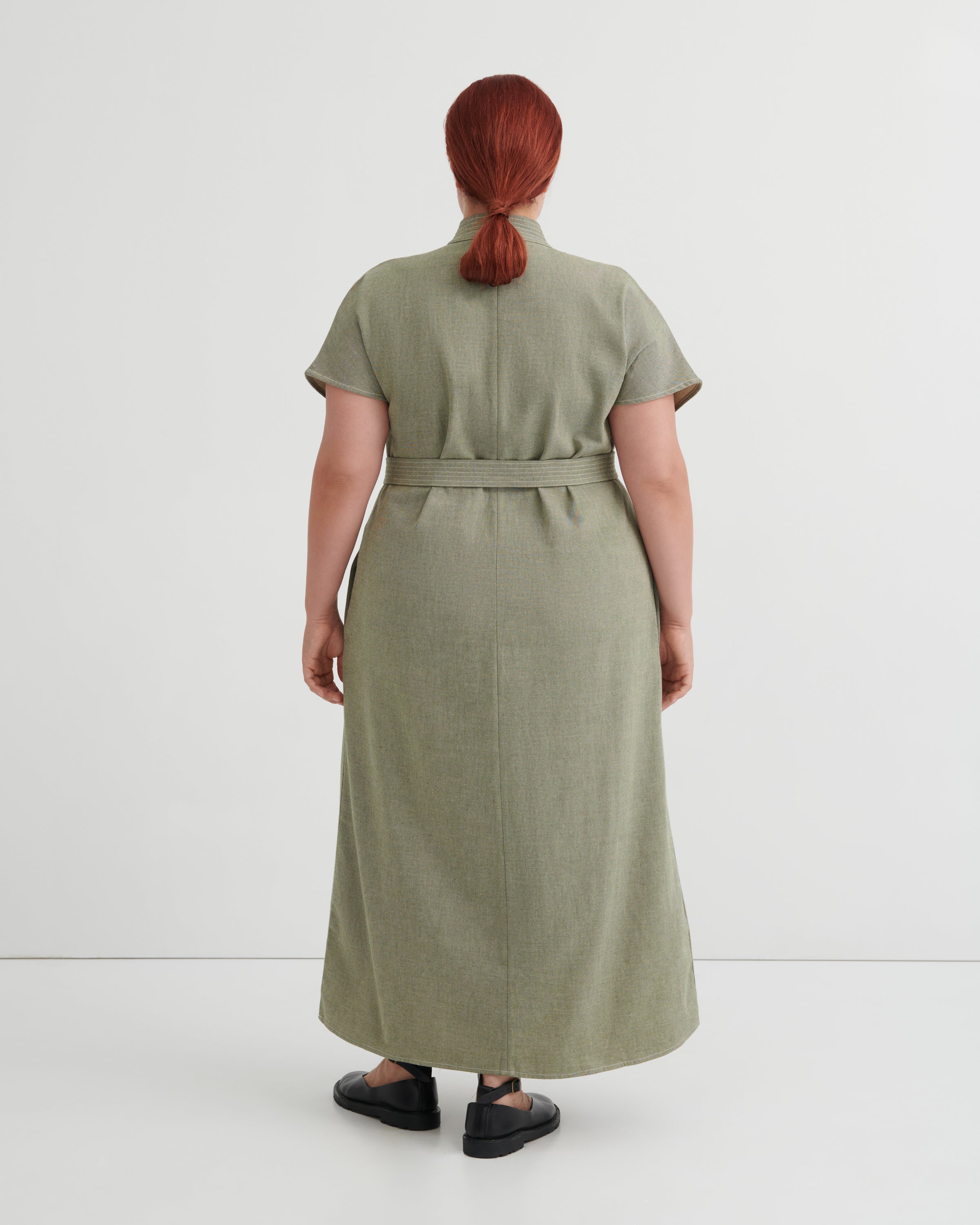 A person shown from the back wearing a short-sleeve, knee-length olive green dress with a matching waist tie, photographed against a clean light gray studio background.