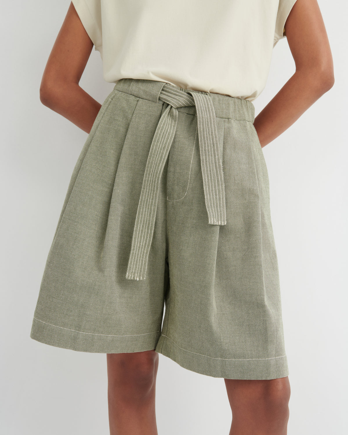 A person wearing high-waisted olive green pleated shorts with a matching tie belt, paired with a cream sleeveless top, against a light gray studio background.