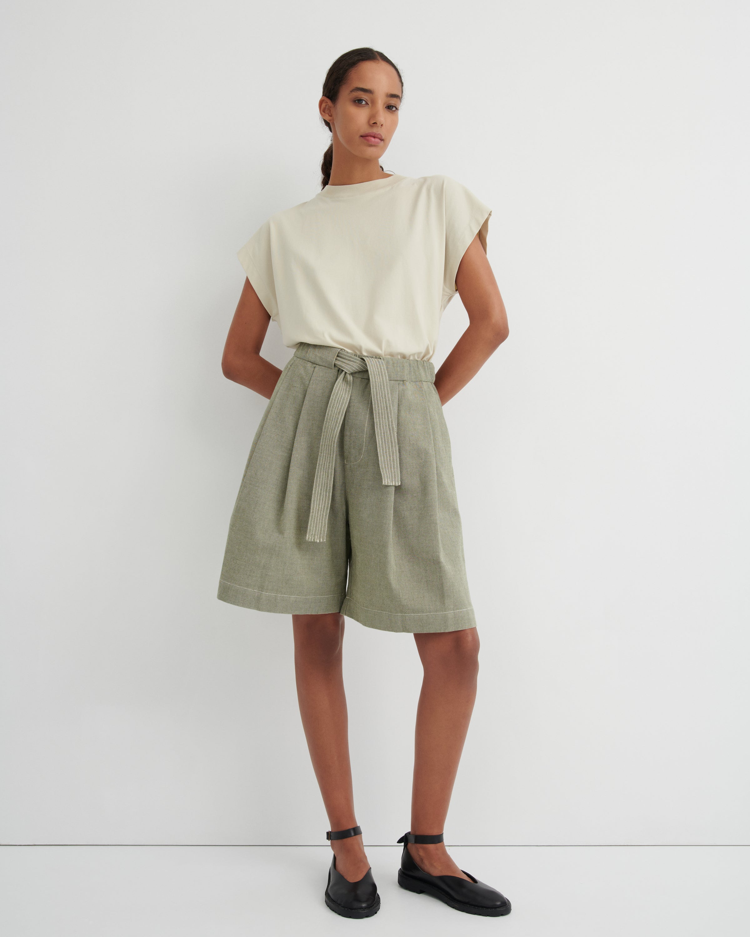 A model stands against a plain white studio wall wearing a cream short-sleeve top and light sage green high-waisted linen shorts with a matching waist tie, paired with black Mary Jane shoes.