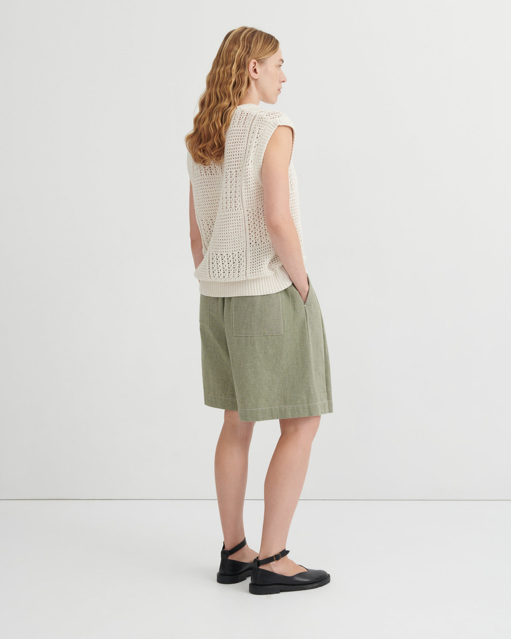 A woman stands in profile wearing a cream sleeveless open-knit sweater vest, sage green shorts, and black Mary Jane flats against a clean white studio background.