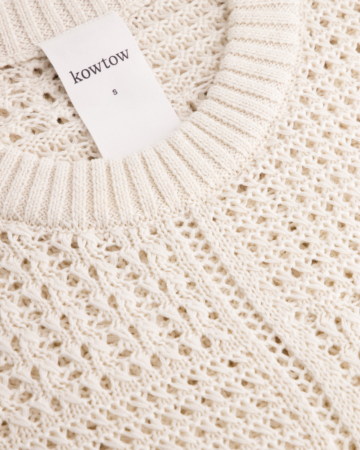 Close-up of a cream-colored knit sweater with a textured openwork pattern and a white brand tag reading 'kowtow' and size 'S'.